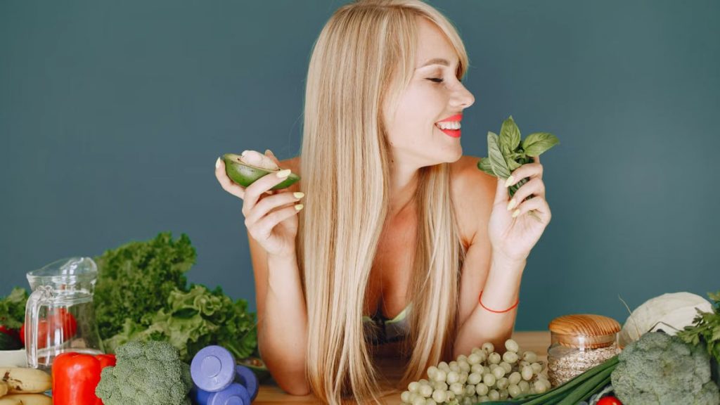 A smiling woman holding avocado and basil, surrounded by fresh vegetables and fruits for healthy eating.